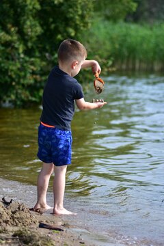 Young boy playing with wet sand and a toy shovel at the edge of a lake on a sunny summer day, enjoying outdoor fun, nature and carefree childhood moments by the water.