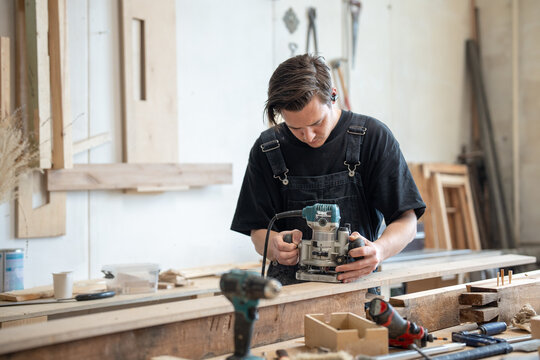 Carpenter restorer working in restoration workshop with electric wood router, man crafter processing wooden board surface with handheld plunge router, craftsman using equipment. Routine in joinery. 