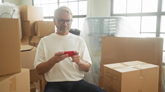 Man holds red smartphone in hand among stacked moving boxes in building, seated by wrapped furniture and large window checking messages; fatigue.