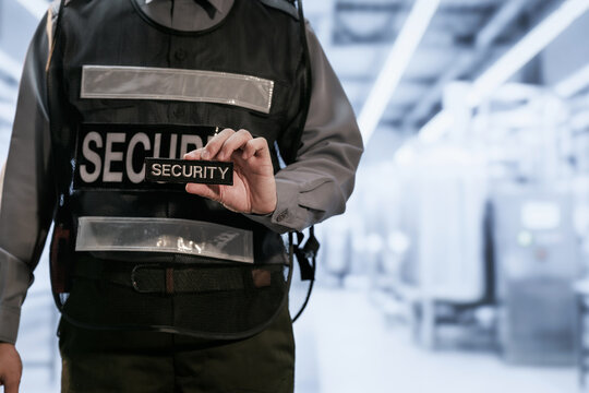 Close-up of a security guard in a black protective vest holding a security badge in a bright modern building lobby.