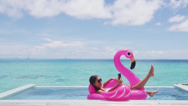Vacation woman on inflatable Flamingo float mattress using phone in swimming pool. Girl relaxing on travel holidays at resort pool in bikini. Raw unedited video of real woman model without makeup