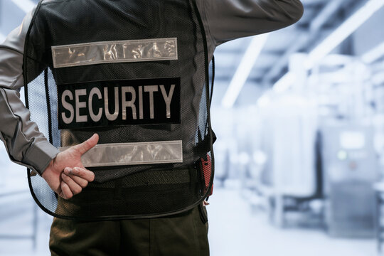 Back view of a security guard in a black vest with a clear security label, standing in a bright modern indoor public space.