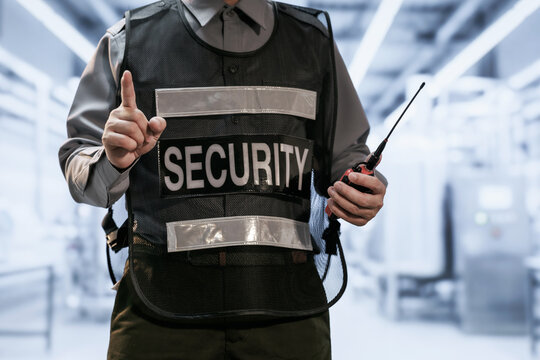 Security guard in a reflective vest holds a radio and raises one finger in a bright modern building entrance.