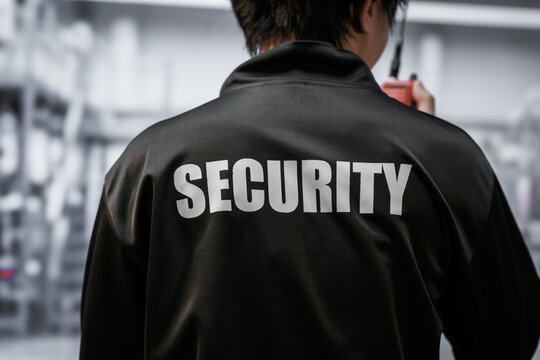 Back view of a security guard in a black jacket with a large security label, standing in a blurred indoor public space.