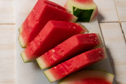 A fresh vibrant ripe red watermelon arranged on a white cutting board. The flesh is bright red and juicy, featuring clearly visible small white seeds and the characteristic green outer rind.
