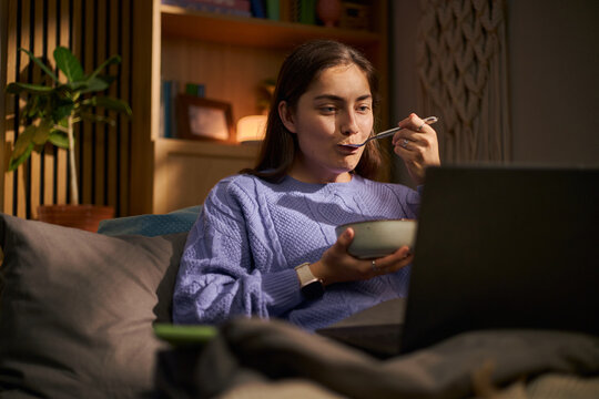 Young adult Caucasian woman eating meal from bowl while watching laptop screen in bed in evening, representing digital lifestyle, suitable for streaming service marketing