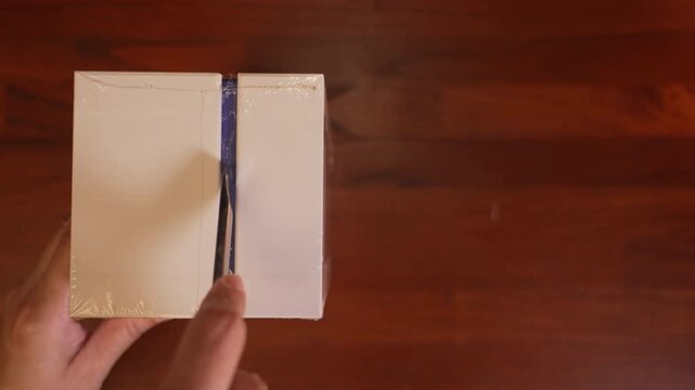 Top view close up of vertical white box on left side leaving copy space, hand slicing protective shrink wrap with utility cutter knife, sealed package delivery concept.