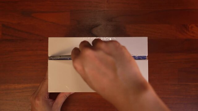 Top view close up of hand using utility knife cutter to slice open plastic shrink wrap on centered white box on wooden desk, e-commerce sealed parcel unboxing concept.