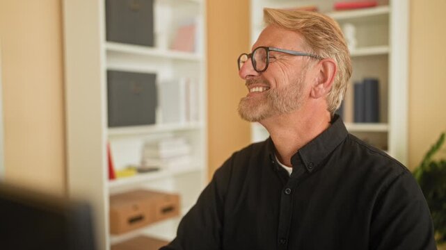 Man at computer smiling and gesturing with hand in building workspace wearing glasses and black shirt, seated near shelves and books; contentment productivity.