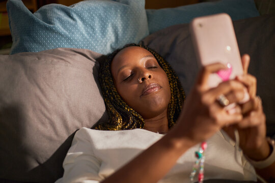 Young adult African American woman lying in bed holding smartphone with closed eyes, representing digital fatigue or sleep issues, ideal for mental health campaigns