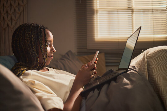 Young adult African American woman using smartphone while lying in bed with laptop, representing remote work and digital lifestyle, suitable for mobile banking or e-commerce ads