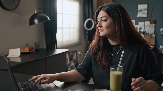 Young adult woman drinking green smoothie and typing on laptop during remote work, camera zooming in. Clip is ideal for wellness apps or remote work lifestyle campaigns