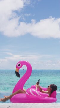 Vacation woman on inflatable Flamingo float mattress using phone in swimming pool relaxing on travel holidays at resort pool in bikini. Raw unedited vertical video of real woman model without makeup