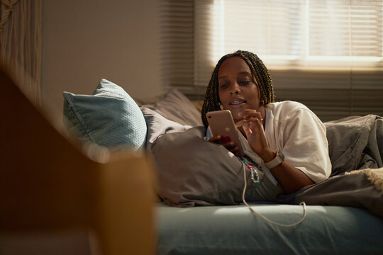 African American girl lying in bed and using smartphone, representing digital life and youth communication, suitable for mobile application marketing
