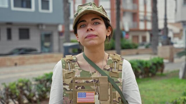 Woman soldier wearing camouflage cap and tactical vest with us flag patch and dog tags looks at camera on street near low wall and palms; pride duty.
