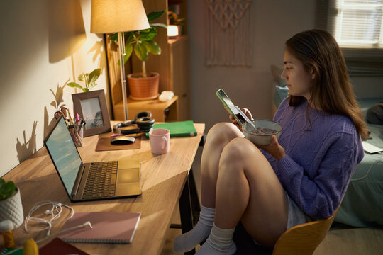 Caucasian girl sitting at desk, eating breakfast and using smartphone, representing digital lifestyle and remote learning, suitable for youth marketing campaigns