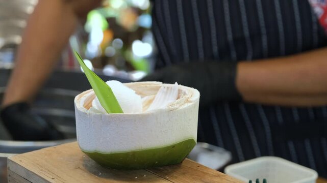 Street vendor assembling a coconut ice cream dessert by carefully placing scoops of ice cream into a hollow coconut shell at a market. Thai street food concept