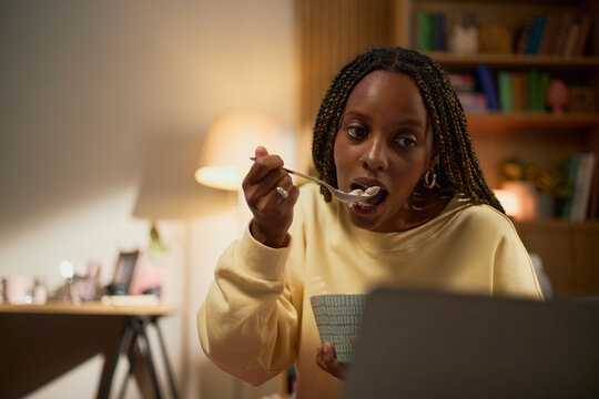 Young adult African American woman eating snack from bowl while watching laptop screen at home, representing remote lifestyle and suitable for digital entertainment marketing