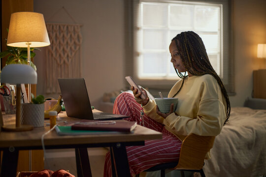 Young adult Black woman sitting at desk in bedroom, using smartphone and holding bowl, representing remote work, digital lifestyle, and leisure, suitable for lifestyle marketing