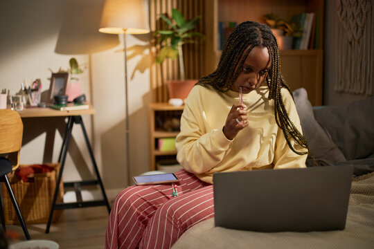 Young adult African American woman sitting on bed, looking at laptop screen and brushing teeth, representing remote learning, online education, and freelance work, medium shot