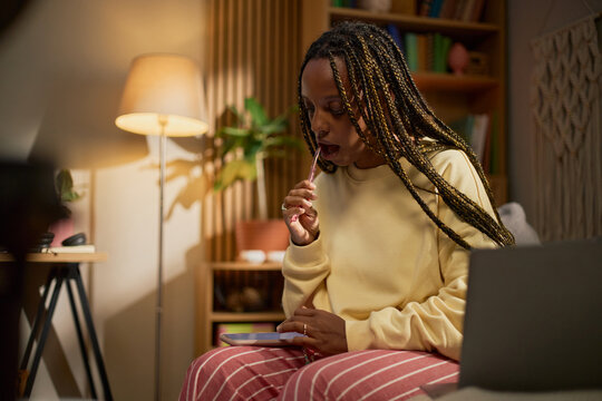 Young adult Black woman using smartphone while brushing teeth at home. Image suitable for remote work, digital lifestyle, or e-learning campaigns