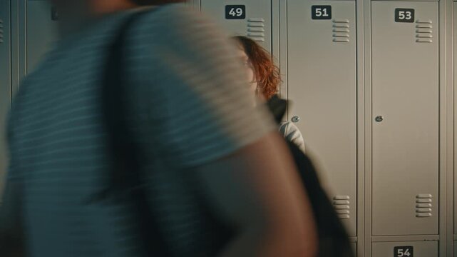 Upset Female Teenager Standing Still by Metal Lockers, Experiencing Sadness and Vulnerability. In Foreground, Classmates Walk Past, Ignoring Presence. Social Indifference, Feeling Invisible in Crowd.
