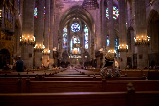 Woman in hat sitting in wooden pew inside gothic cathedral interior with stained glass windows and candles in Mallorca, religious architecture scene