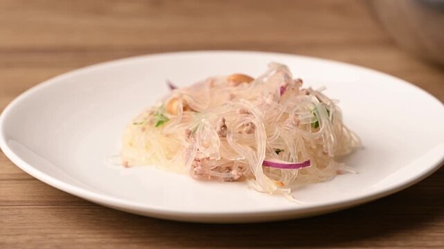 Close-up of tongs plating Thai spicy glass noodle salad (Yum Woon Sen) onto a white plate, preparing for serving