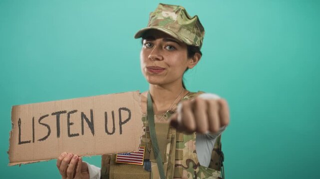 Woman soldier holding a cardboard sign reading listen up with fist extended toward camera in studio; authority call to action.