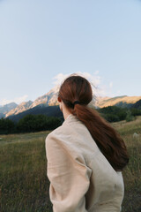 A woman stands in an open field with distant mountains, wearing a light jacket, hair tied back. A lifestyle scene of calm outdoor reflection and natural beauty. © SHOTPRIME STUDIO