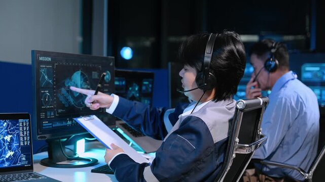 Asian engineer in headset analyzes radar screens and takes notes in a high-tech control room, managing telemetry
