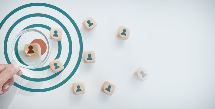 Hand holding magnifying glass focusing on target bullseye with selected wooden block human icon among group, concept of target audience, recruitment, and human resource management, Niche Marketing.
