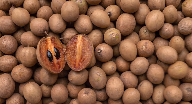 Halved fresh sapodilla (sawo / chiku / chickoo) revealing brown flesh and black seeds surrounded by a pile of whole ripe sapodillas in full frame layout.