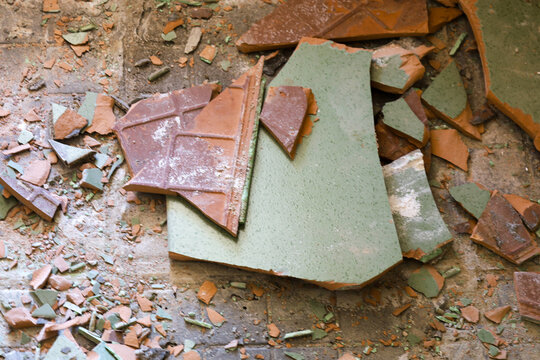 A pile of broken old ceramic tiles lies on the floor after demolition. A chaotic mix of green and terracotta fragments creates a raw texture of construction debris. Remnants of the concrete base are v