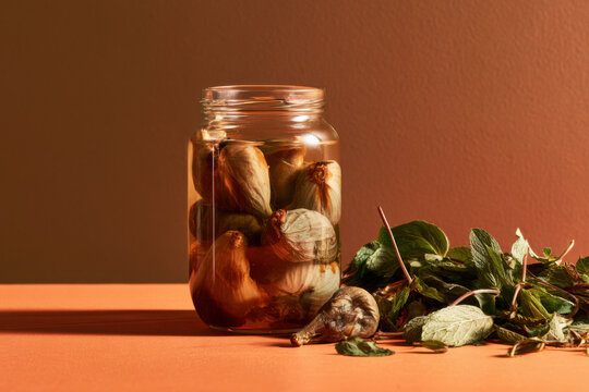 Rustic glass jar filled with preserved garlic bulbs in liquid beside fresh herbs, styled with warm earthy lighting and generous copy space.