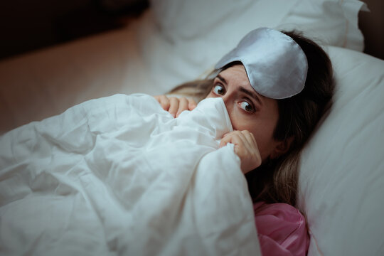 Shy Scared Woman Hiding Under White Blanket Wearing Silk Sleep Mask In Bed. Expressive young woman peering out from under duvet with surprised or timid look