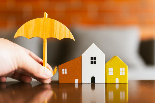 Property insurance and real estate protection concept with hand holding wooden umbrella over miniature house models on table with warm brick background for mortgage safety and coverage.