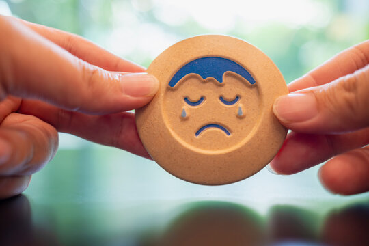 Mental health and depression concept with hands holding wooden token showing sad crying face emoji on green blurred background for psychological support, sorrow and negative feedback.