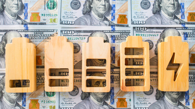 Energy cost and electricity bill concept with wooden battery charge level icons on hundred dollar bills for power consumption expense, renewable investment and green storage finance.