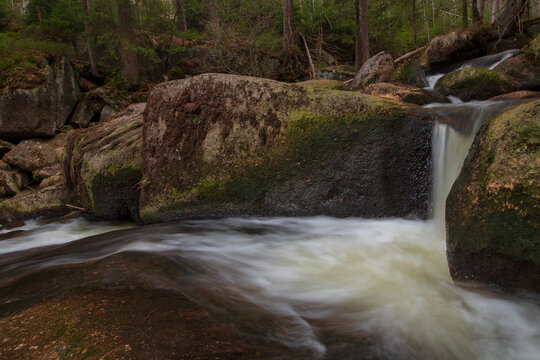 Mountain stream flows through massive moss-covered rocks in a forest in the Jizera Mountains, Czech Republic, captured with a slow shutter speed.