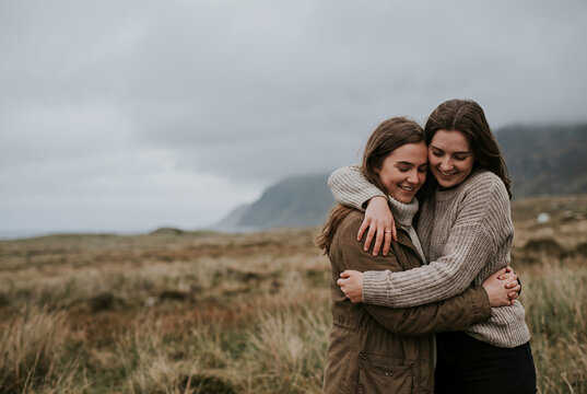 Two young women embracing outdoors with mountain landscape and overcast sky