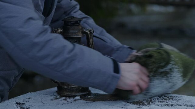 Person using a folded blanket to clear snow off a wooden table beside a burning vintage lantern