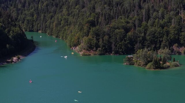 Small boats glide across a vivid turquoise lake surrounded by a dense, verdant forest under clear daylight.