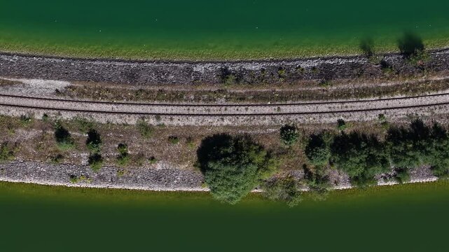 An aerial view shows parallel train tracks running alongside a rocky embankment and serene green water.