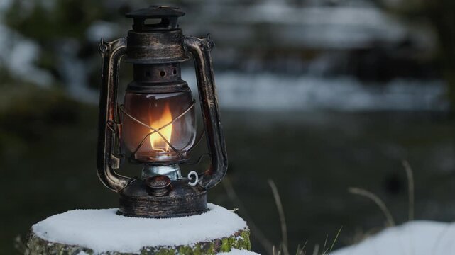 Close-up of a glowing vintage lantern on a snowy tree stump with a blurred flowing stream in the background