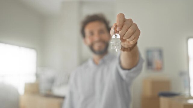 Man outstretched hand holding house key among moving boxes in a building interior with blurred window; new home joy.
