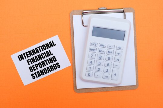 A white calculator on a clipboard next to a white note with text "INTERNATIONAL FINANCIAL REPORTING STANDARDS". Concepts of accounting, IFRS compliance, global finance and business auditing