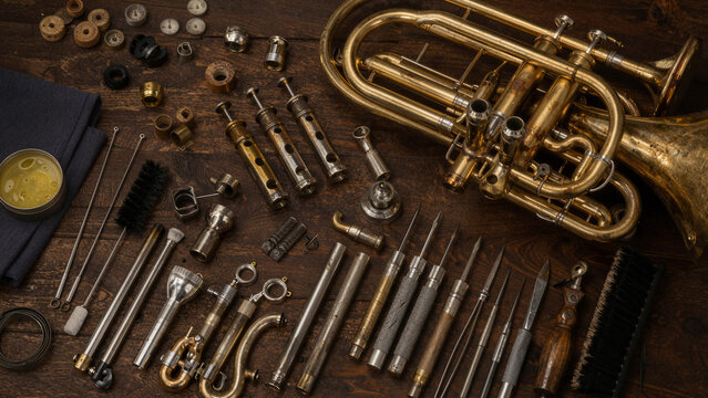 Several brass instrument repair tools and parts are displayed on a wooden table. Items include valves, brushes, and cleaning supplies. The lighting is soft, revealing details of metal surfaces