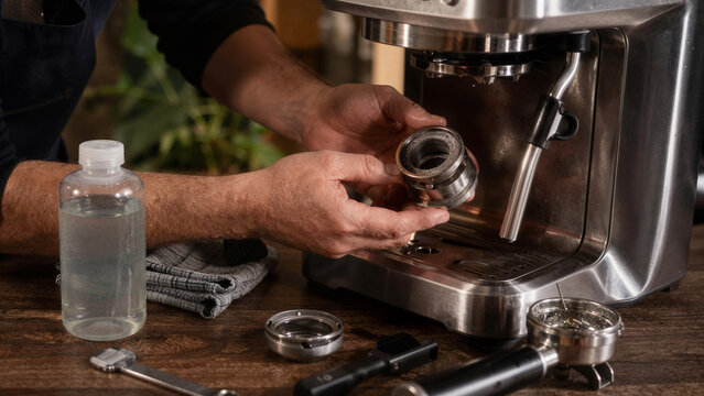A person works in a coffee shop handling a part of an espresso machine. They use a bottle of cleaning solution and tools on the table. It is midday