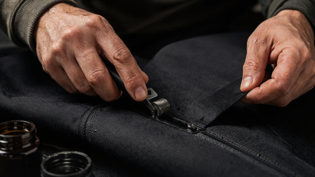 A person uses a tool to fix a zipper on a wetsuit. The setting is a workshop with supplies like glue nearby. The focus is on the hands and the wetsuit